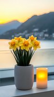 Yellow daffodils in textured gray vase with glowing candle on windowsill at sunset, mountain silhouettes and lake view creating peaceful atmosphere.