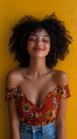 Young African American woman with natural curly hair smiling with eyes closed against yellow wall, wearing floral off-shoulder top and denim, radiating joy and confidence.