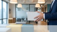 Professional businessman in navy suit holding blank white tablet in modern office conference room with glass walls and pendant lighting for presentation mockup.