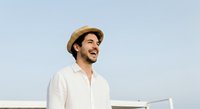 Man in straw hat and white shirt smiling outdoors against clear sky. Young adult male, Mediterranean ethnicity, casual summer style, suitable for lifestyle and fashion content.