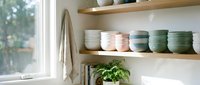 Organized kitchen shelves displaying stacked ceramic bowls in neutral and sage tones, potted green plant, and folded linens near bright window for home decor and interior design.