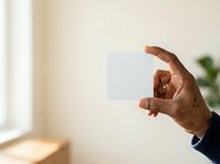 Male hand with dark skin tone holding blank white card or paper against neutral beige wall background, suitable for business presentations and marketing materials.