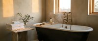 Luxurious spa bathroom with freestanding black bathtub, golden fixtures, natural light from windows, towels, plants, and neutral earth tones for wellness and relaxation design projects.