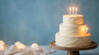 Three-tier white wedding cake with lit candles on wooden stand against blue background for celebration and special occasions.