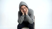 Young Asian man in gray hoodie sitting on chair holding head in hands expressing stress, depression, anxiety, mental health struggles, emotional pain.