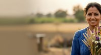 Portrait of a middle-aged South Asian woman wearing traditional blue attire with gold embroidery and a bindi, smiling outdoors near agricultural fields and water. Suitable for cultural diversity, farming communities, and lifestyle content.