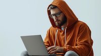 Young bearded man in orange hoodie and glasses working on laptop computer against light background, modern remote work lifestyle concept.