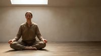 Calm middle-aged Caucasian man practicing meditation in lotus position on wooden floor in minimalist room with natural skylight for wellness and mindfulness.