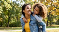 Happy African American mother carrying daughter on back outdoors in autumn park, family bonding moment with golden sunlight and trees.
