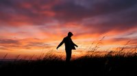 Woman silhouette standing in tall grass field against dramatic sunset sky with orange and purple clouds creating peaceful moment of freedom and contemplation.