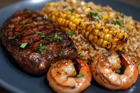 Grilled steak and shrimp surf and turf dinner served with roasted corn on cob and brown rice, garnished with fresh herbs on dark plate.