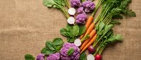 Fresh organic vegetables including purple cauliflower, carrots, radishes and leafy greens arranged on natural burlap background for healthy cooking.