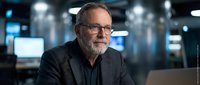 Mature male professional with gray beard and glasses working in modern tech office environment with laptop and blue-lit background for business and corporate concepts.
