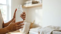 Young adult with medium brown skin tone holding white cosmetic or cleaning product bottle in bright bedroom environment, promoting wellness and home care.