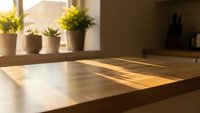 Modern kitchen countertop with warm sunlight streaming through window creating natural shadows on wooden surface with potted plants in background.