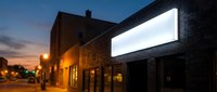 Large blank white billboard mounted on brick building wall during twilight hour with warm street lighting creating urban advertising space for marketing campaigns.