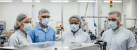 Four diverse medical professionals in surgical masks and hair nets standing together in modern sterile laboratory or cleanroom facility for teamwork.