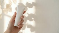 Caucasian woman's hand holding blank white cosmetic bottle with natural sunlight casting shadows on neutral background for skincare product mockup.