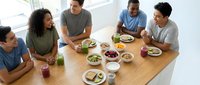 Diverse group of young adults enjoying healthy meal together around wooden dining table in bright modern kitchen sharing food and conversation.