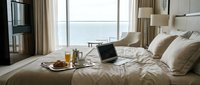 Luxury hotel bedroom with laptop on bed, breakfast tray, ocean view through large windows. Modern hospitality interior for business travel and remote work.