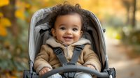 Cheerful mixed race baby in warm winter clothes sitting in grey stroller outdoors during autumn season, smiling at camera against blurred yellow foliage background.