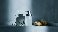 Minimalist spa bathroom setup with pump dispenser bottle, natural sponges and eucalyptus leaves on marble surface against textured blue wall for wellness branding.