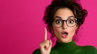 Young woman with surprised expression wearing glasses and green turtleneck sweater raises index finger against vibrant pink background, showing eureka moment.