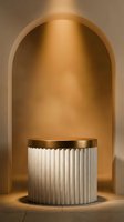 Minimalist cylindrical pedestal with fluted white base and golden top in warm beige archway niche for luxury product display and modern interior design.