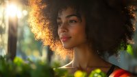 Young African American woman with natural afro hair illuminated by golden sunlight, smiling confidently in nature setting with soft bokeh background and warm sunset glow.