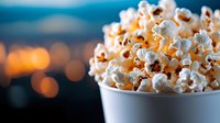 Fresh popcorn overflowing in white paper container against blurred city lights background, warm evening atmosphere creates cozy movie time ambiance.