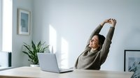 Young Asian woman stretching arms above head while working at laptop in bright modern office space with plants and natural light for wellness breaks.