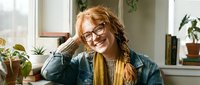 Young blonde woman with glasses smiling warmly while resting chin on hand in bright home office with plants. Casual denim jacket and cozy indoor lifestyle setting.