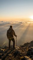 Hiker with backpack and trekking pole standing on mountain peak above clouds at golden sunrise, enjoying panoramic view of landscape below.