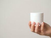 Woman's hand holding white cosmetic jar with manicured nails against neutral background, suitable for skincare, beauty product packaging, wellness advertising, and cosmetics marketing campaigns.