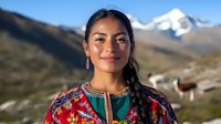 Young Latin American woman in traditional colorful embroidered dress smiling outdoors against mountain landscape with snow peaks and llama in background.