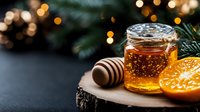 Glass jar of golden honey with wooden dipper and fresh orange slice on rustic wood slice against dark background with pine branches and bokeh lights.