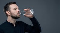 Adult male with dark hair and beard, Caucasian ethnicity, drinking water from bottle against gray background. Suitable for health, fitness, wellness and hydration lifestyle content.