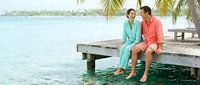 Couple relaxing on wooden dock over turquoise water at tropical resort. Man and woman in colorful casual wear enjoying romantic vacation by ocean pier.