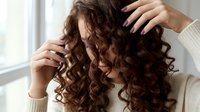 Young woman with beautiful curly brown hair running fingers through locks in natural light by window. Hair care routine and styling concept.