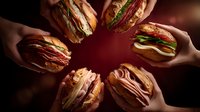 Multiple hands holding gourmet sandwiches in circular arrangement on dark background. Fresh deli meat, cheese, lettuce creating appetizing fast food display for sharing.