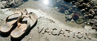 Flip flops on sandy beach with word vacation written in pebbles near coral rocks and sparkling sea water on sunny summer day.