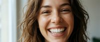 Happy young woman with curly brown hair smiling broadly showing white teeth in bright natural light indoor setting for lifestyle and wellness content.