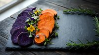 Sliced purple and orange vegetables arranged on dark slate board with microgreens and herbs for gourmet cooking and healthy meal preparation.