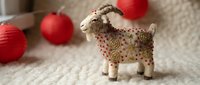 Young goat figurine with spotted brown and cream pattern standing on neutral textile surface with red ornaments in soft focus background, suitable for holiday decorations, seasonal marketing, and festive design projects.