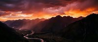 Dramatic mountain landscape with winding river at sunset, golden and crimson sky with storm clouds, silhouetted peaks, and glowing valley below for nature and travel projects.