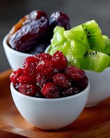 Fresh dried cranberries, sliced kiwi fruit, and dates arranged in white ceramic bowls on wooden surface, healthy snack food composition in natural light.