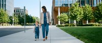 Young mother and toddler son walking hand in hand on urban sidewalk in modern business district with glass buildings and green spaces for family lifestyle.