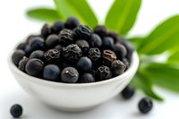 Fresh black acai berries in white ceramic bowl with green leaves on white background, macro shot for healthy food and superfood marketing.