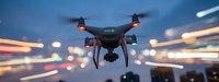 Black quadcopter drone flying over blurred city lights at dusk with colorful bokeh background for aerial photography and videography projects.