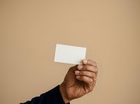 Male hand with dark skin tone holding blank white business card against neutral beige background for corporate branding and professional networking purposes.